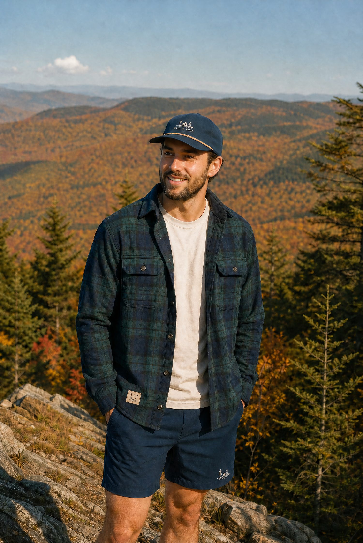 A man in a navy plaid flannel and Salt & Pine cap on a mountain overlook above autumn foliage